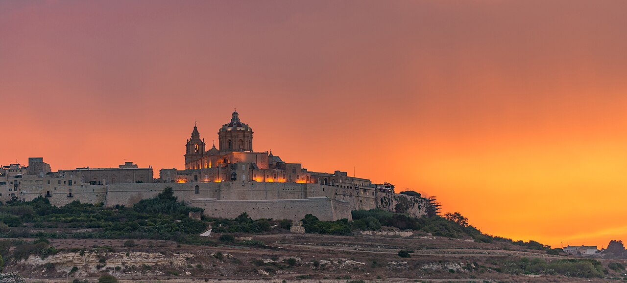 Mdina’s fortified hilltop and cathedral silhouetted against an orange sunset.