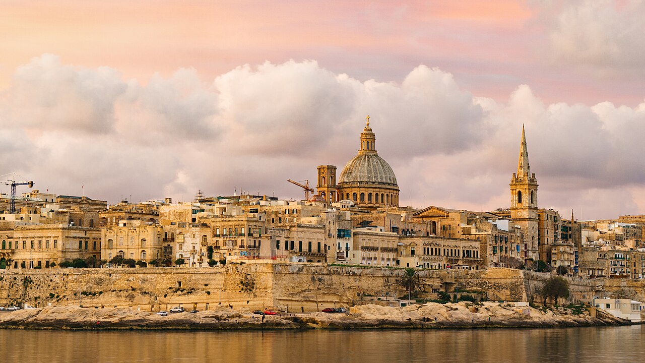 Valletta’s limestone waterfront, Carmelite dome and church spire beneath a pink evening sky.